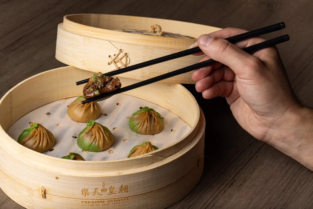 A hand lifting a chocolate xiao long bao from a steamer of green and brown dumplings